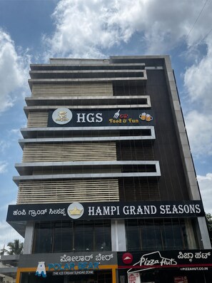 Un edificio de varios pisos con diversos establecimientos comerciales en la planta baja, incluyendo un restaurante llamado 'HAMPI GRAND SEASONS' y una pizzería.