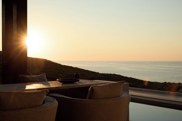 A table with chairs and a bowl, overlooking a coastal view with the sun setting.