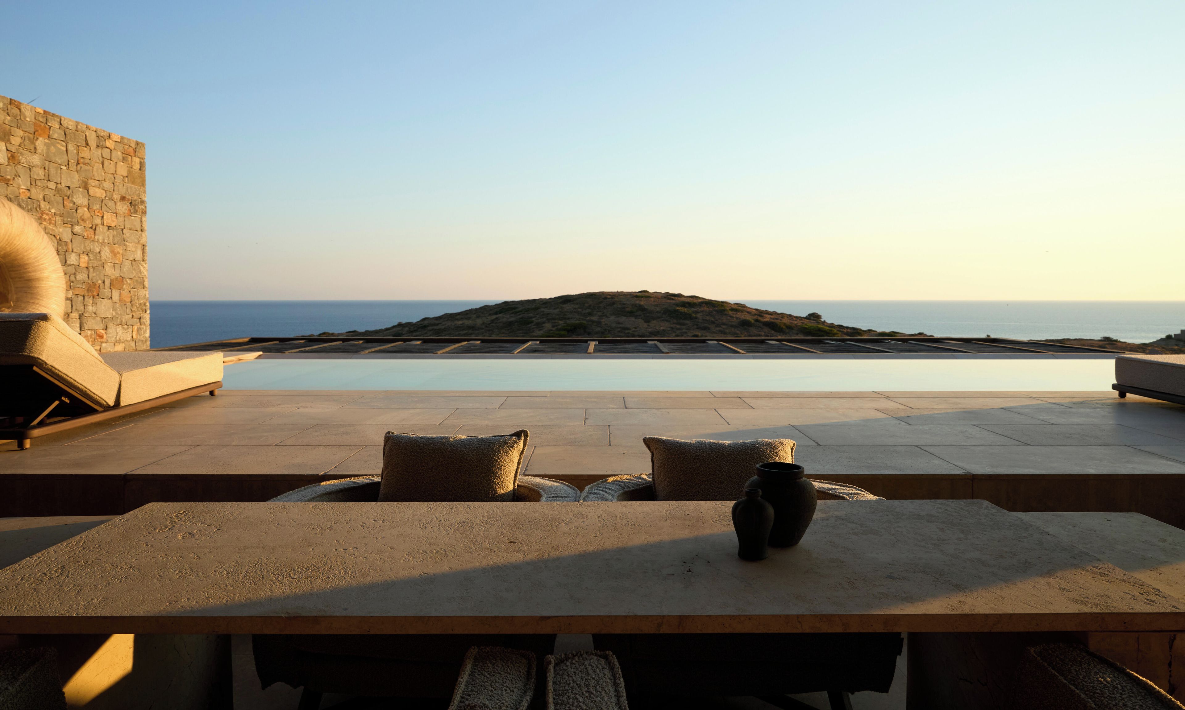 Zona junto a la piscina con vista al mar, pared de piedra y mesa con cojines.