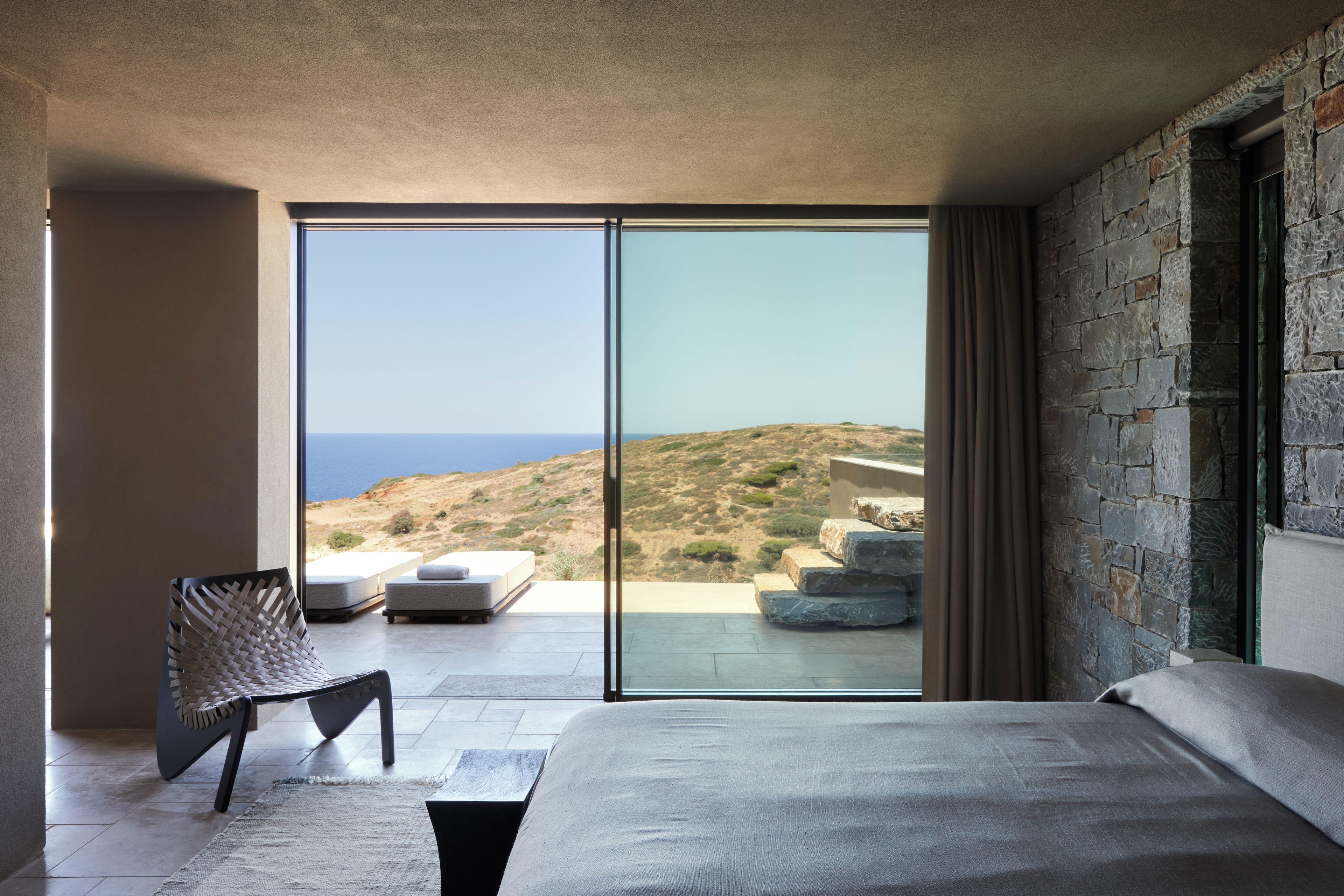 Un dormitorio moderno con una cama grande, un área de descanso con una silla y vistas a un paisaje costero a través de ventanas de piso a techo.