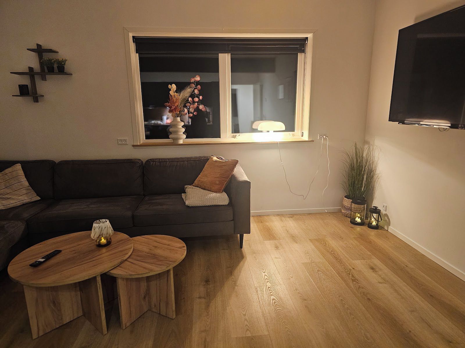 A living room with a sofa, two round wooden tables, a window with a plant on the sill, a TV mounted on the wall, and a wooden floor.