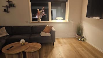 A living room with a sofa, two round wooden tables, a window with a plant on the sill, a TV mounted on the wall, and a wooden floor.