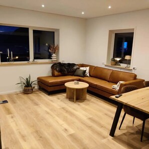 A modern living room with a brown leather sofa, a round wooden coffee table, and a dining table with chairs. There are two windows with curtains, a potted plant, and a small table with decorative items.