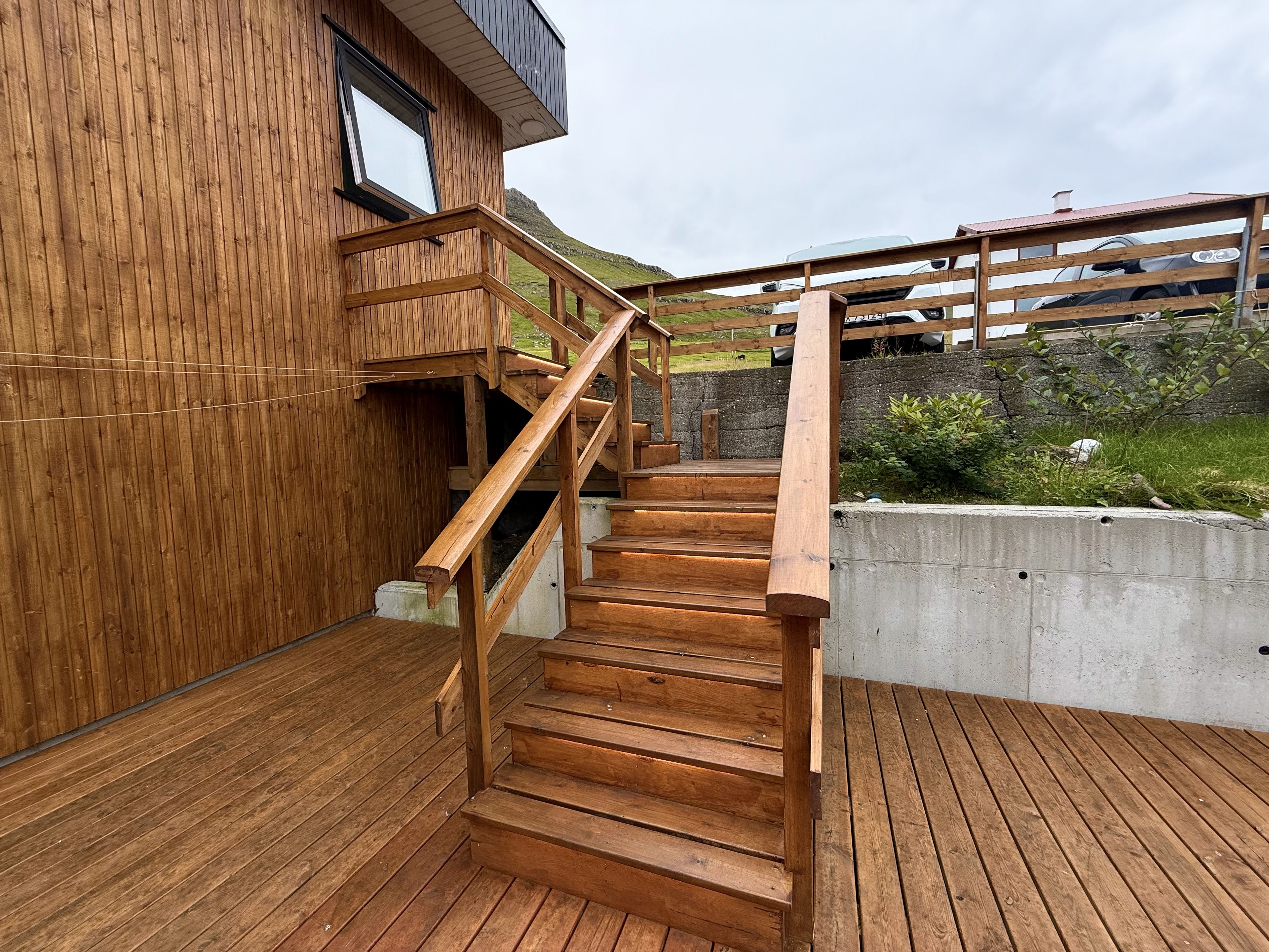 Una escalera de madera que conduce a un edificio con balcón, rodeado de follaje y un muro de piedra.