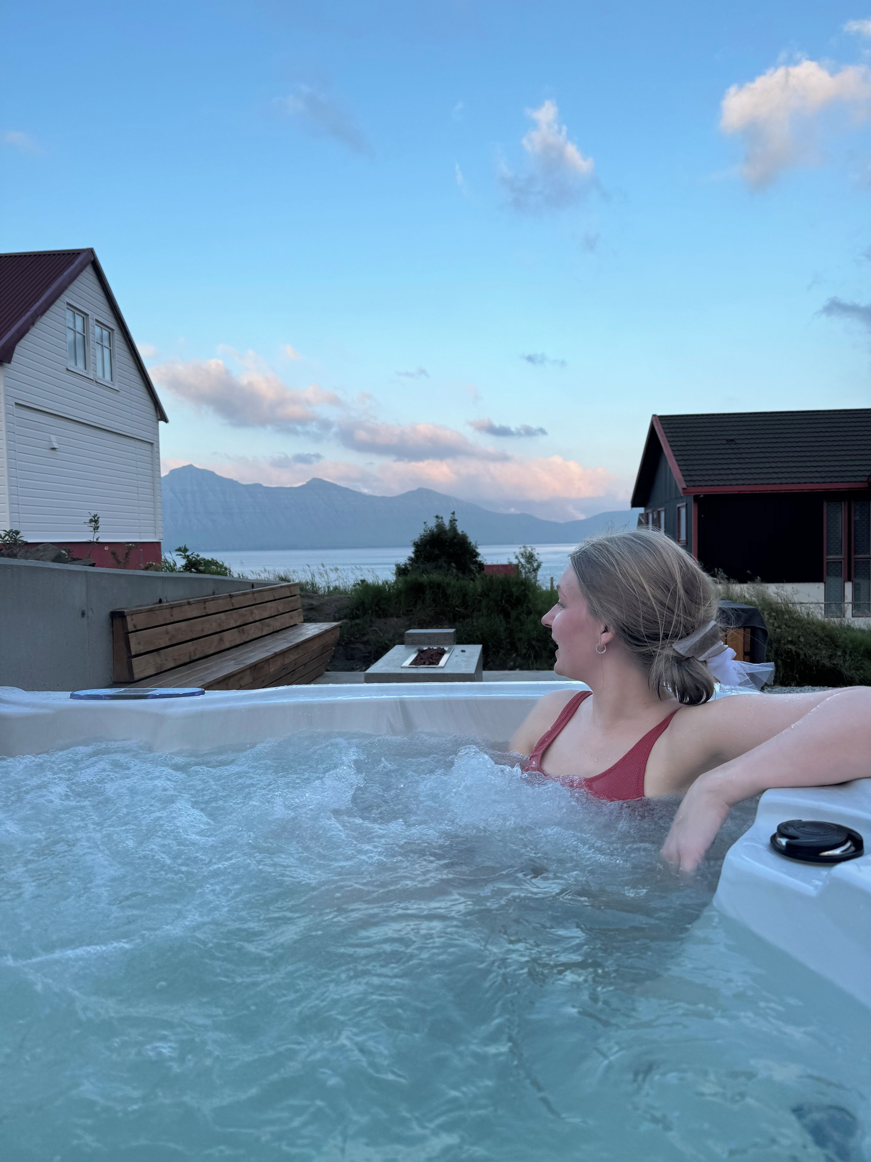 Un jacuzzi con una persona adentro, rodeado por una terraza de madera y vistas a montañas y un lago.