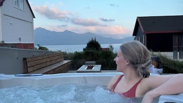 Un jacuzzi con una persona adentro, rodeado por una terraza de madera y vistas a montañas y un lago.