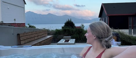 Un jacuzzi con una persona adentro, rodeado por una terraza de madera y vistas a montañas y un lago.