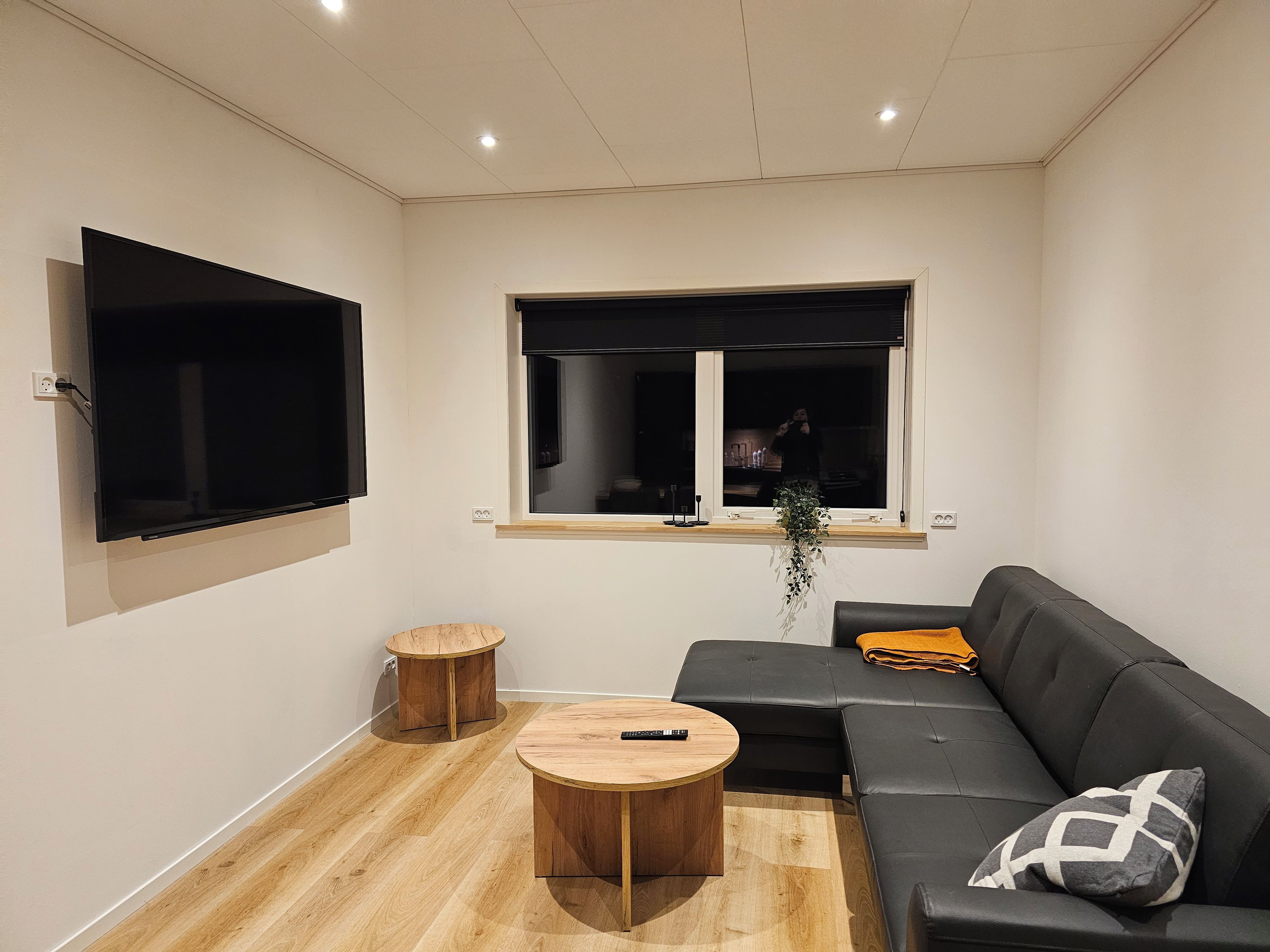 A modern living room with a flat-screen TV, a wooden coffee table, a black sofa, and a window with a view of the outside.