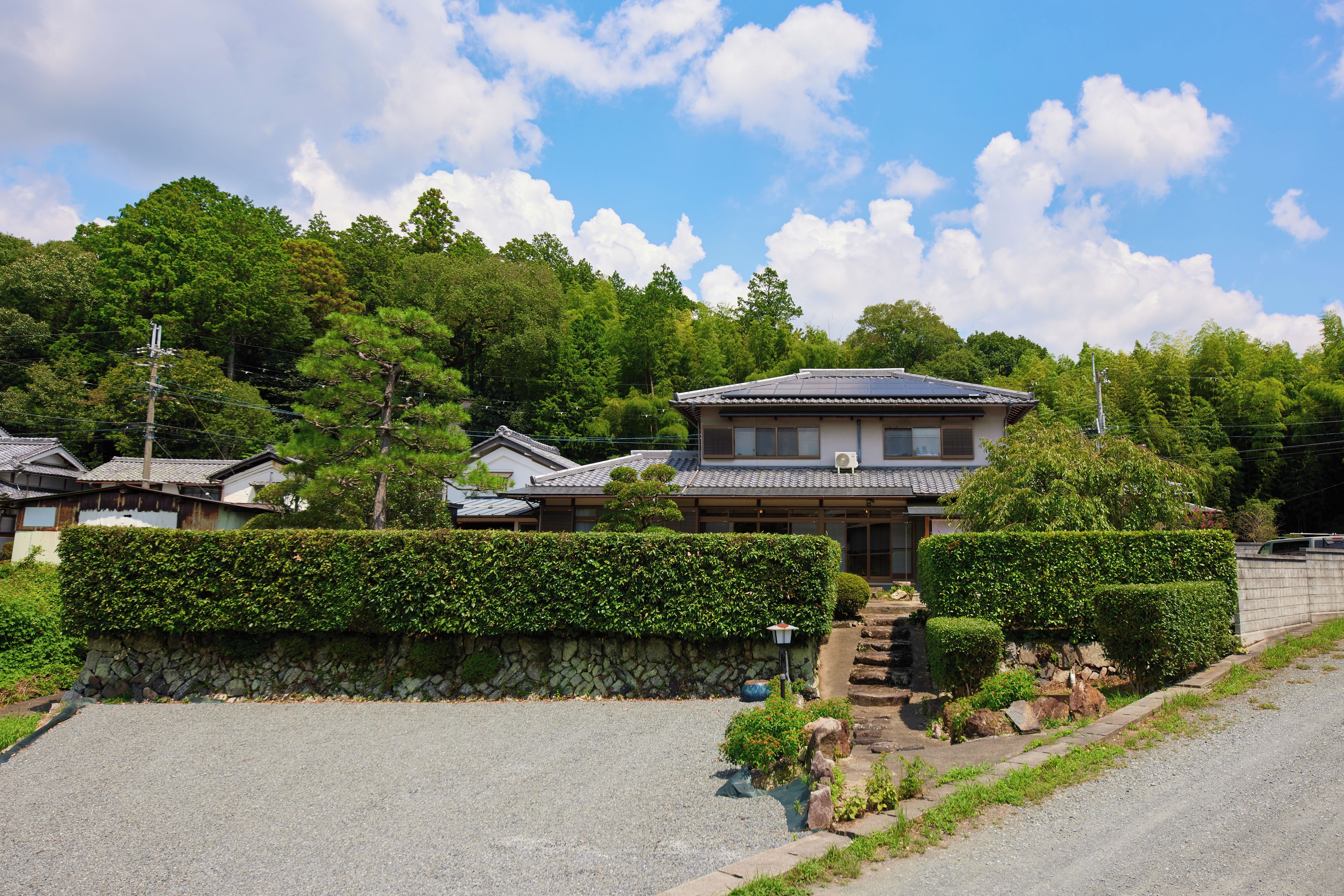 A traditional Japanese house with a tiled roof, surrounded by lush greenery and a stone wall.