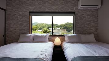A hotel room with two beds, a window with a view of buildings and greenery, and an air conditioning unit on the wall.