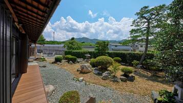 A traditional Japanese garden with a wooden deck, rock formations, and a view of distant houses and mountains.