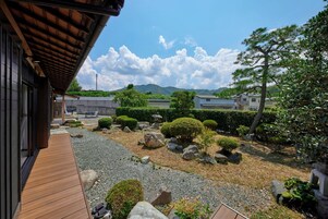 Un jardín japonés tradicional con un suelo de madera, formaciones rocosas y vistas a casas y montañas distantes.