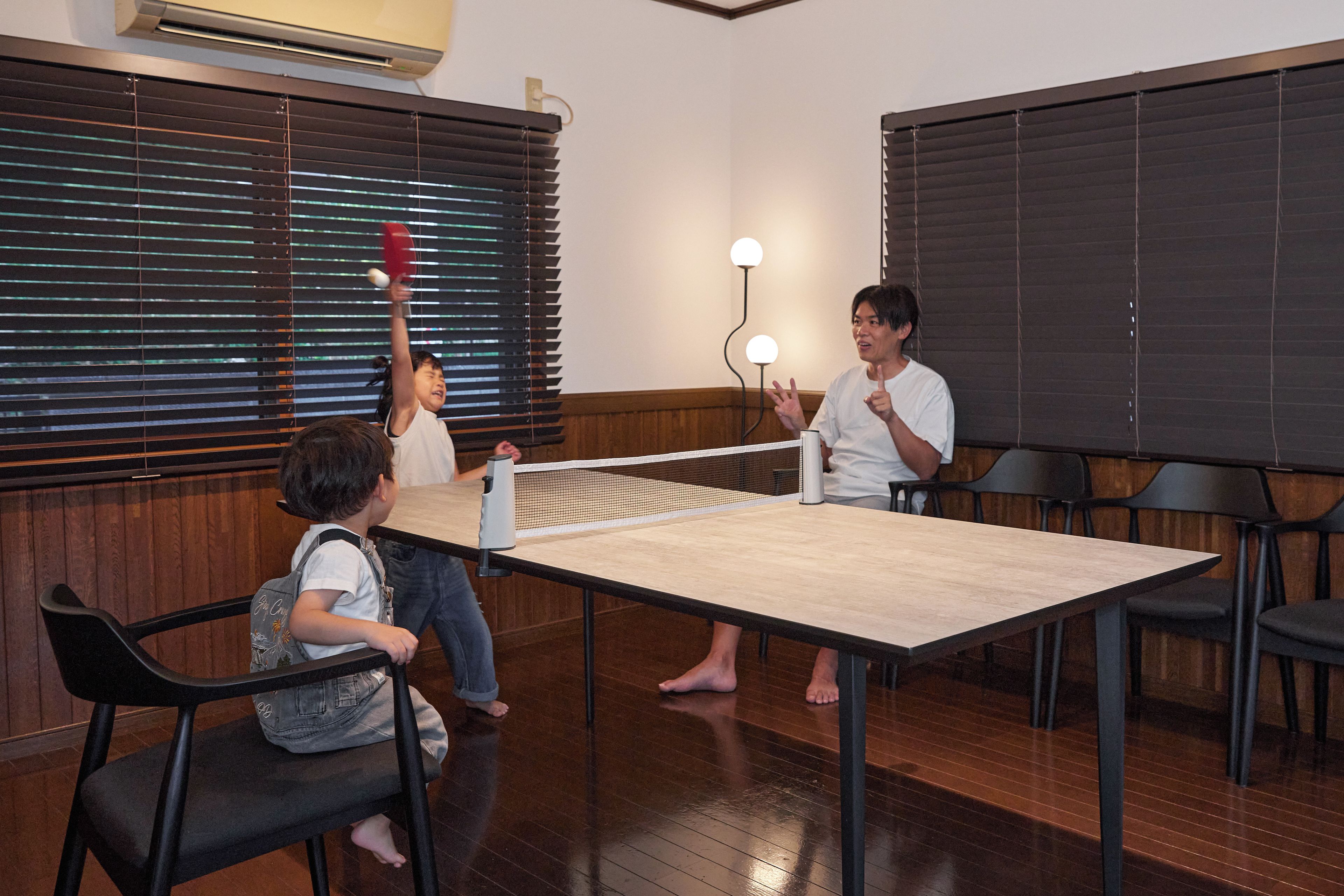 Una habitación con mesa de ping pong, piso de madera y una ventana con persianas.
