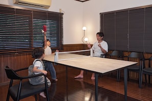 Una habitación con mesa de ping pong, piso de madera y una ventana con persianas.