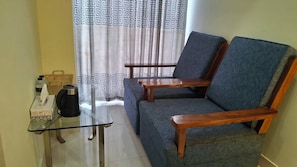 A living area with a sofa, two wooden chairs, a glass table, a kettle, and a basket.
