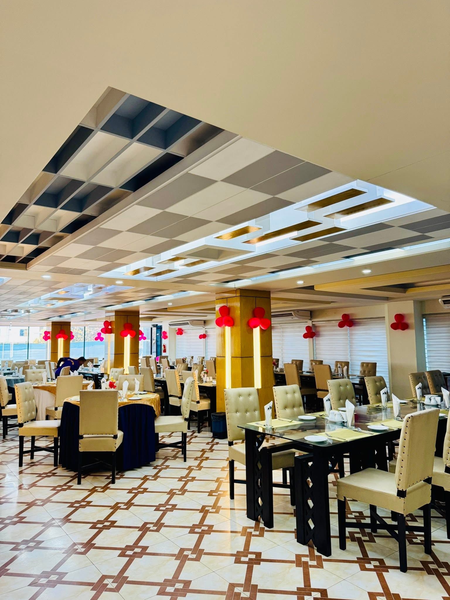 A spacious dining area with multiple tables and chairs, a patterned floor, and a ceiling with a geometric design.