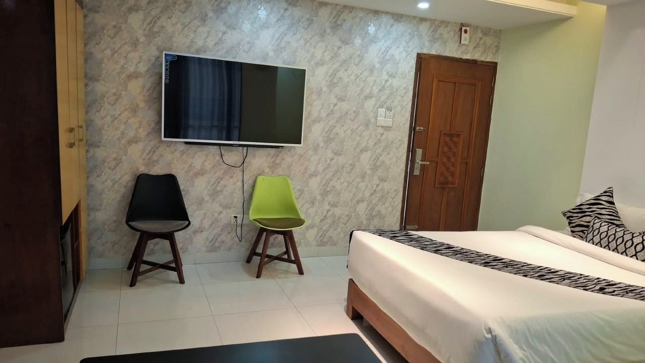 A hotel room with a bed, a TV mounted on the wall, a wooden door, and two chairs.