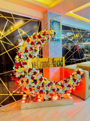 A reception area with a floral arrangement forming the shape of a grand piano, a sign reading 'The Grand Place,' and a modern interior design with geometric patterns on the walls.