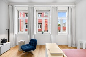 Ein modernes Zimmer mit blauem Sessel, weißem Schreibtisch und einem Bett. Drei Fenster mit Vorhängen bieten Blick auf rote Gebäude.