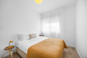 A minimalist bedroom with a bed, a bedside table, a lamp, and a window with sheer curtains. - Bright Apartment | Trindade & City Centre (Porto)