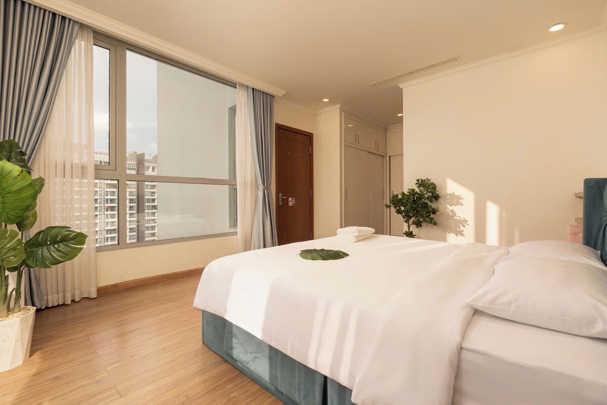Ein Hotelzimmer mit großem Fenster, einem Bett mit weißen Leinen, einer Topfpflanze und Blick auf Gebäude.