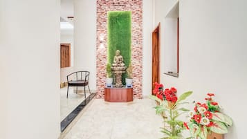 Un pasillo con pared de ladrillo, estatua de Buda, una silla de madera y plantas en macetas.