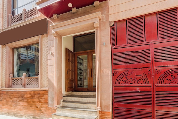 Une entrée d’immeuble avec une porte rouge, un escalier menant à l’entrée et une fenêtre ornée d’un motif en croisillons.