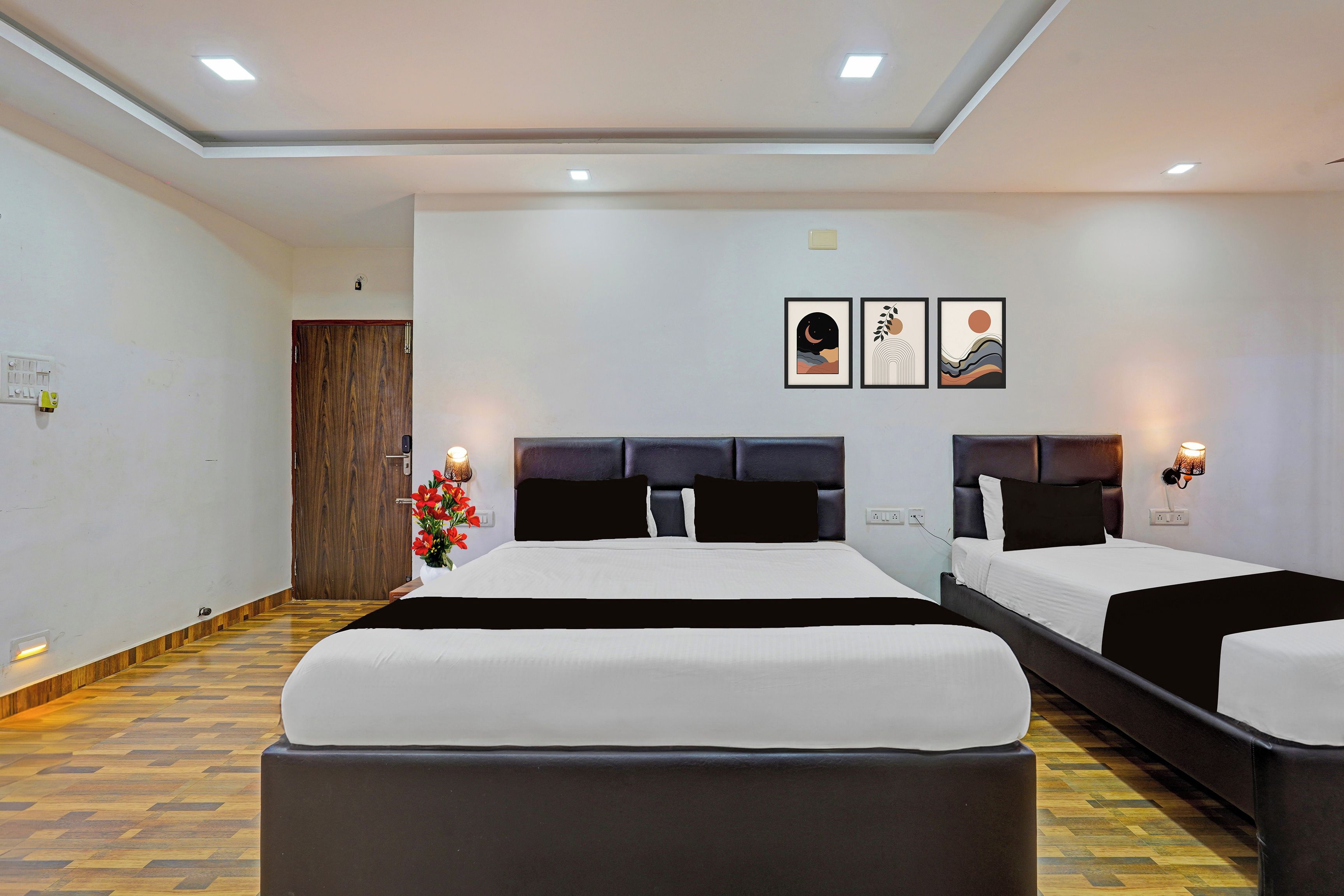 A hotel room with two beds, a wooden door, a flower arrangement, and three framed artworks on the wall.
