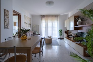 A modern living room with a dining area, featuring a wooden table, chairs, a coffee table, and a sofa. There are potted plants, a TV, and a bookshelf. - A bright 75-square-meter three-room apartment, for up to six guests, located on the first floor of a building (equipped with an elevator, though not suitable for people with disabilities).It enjoys a strategic location near Veronafiere and offers excelle (Verona)