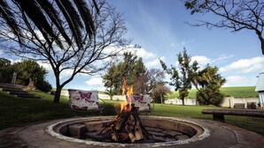 A circular fire pit with burning logs, surrounded by a grassy area with trees and a bench. - Dassenheuwel Farm Stay Homestead (Malmesbury)