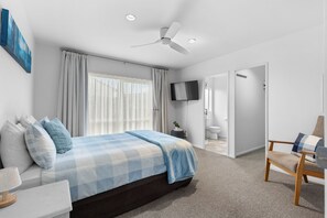 Ein modernes Schlafzimmer mit einem großen Bett, einem Deckenventilator, einem Fernseher und einem durch eine offene Tür sichtbaren Badezimmer.