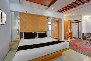 A modern bedroom with a bed, a wooden headboard, a mirror, a sliding door, and a small seating area with a chair and table. - Classic Hotel Vrundavan Residency (Vadodara)
