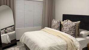 A bedroom with a bed, a bedside mirror, a window with blinds, and a rug. - Apartment in Stay Near Montecasino (Johannesburg)