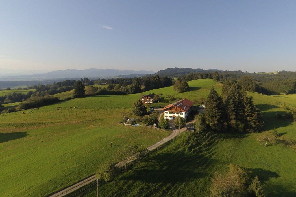 A rural property with a house surrounded by green fields and trees, a clear sky, and distant mountains.