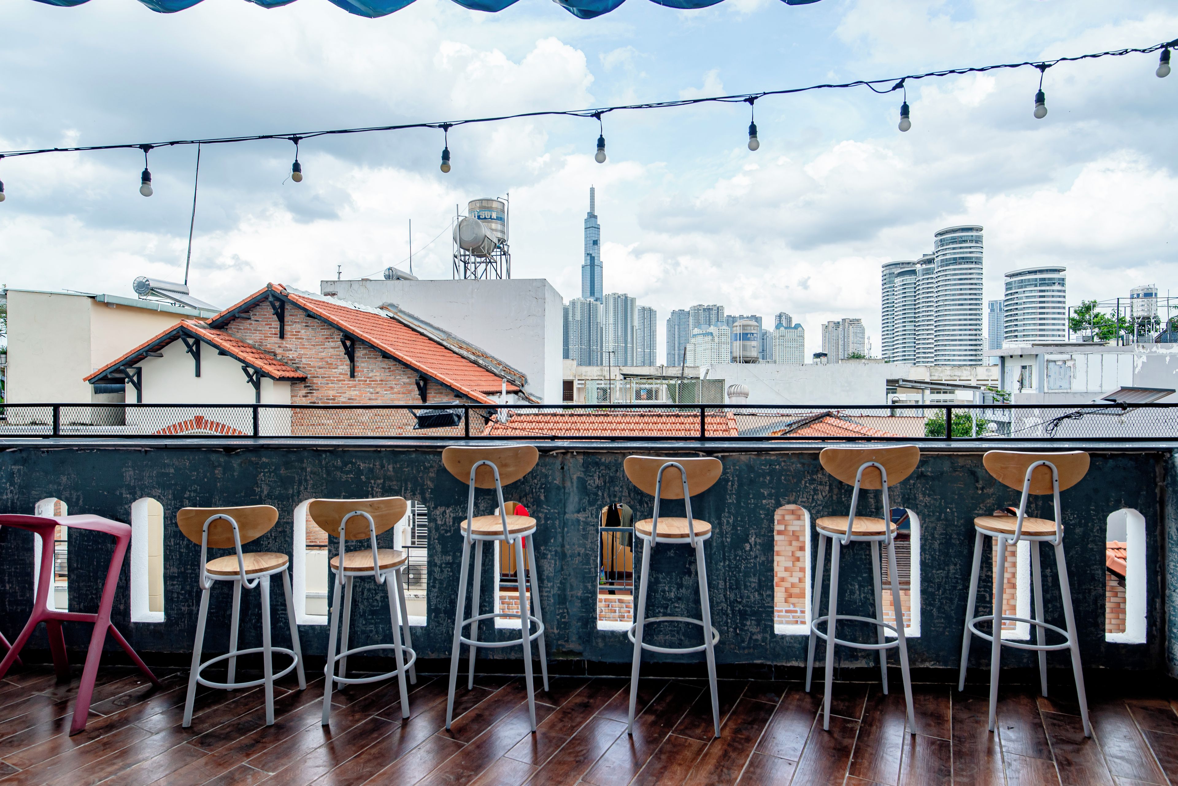 A rooftop bar with wooden flooring and metal bar stools, overlooking a city skyline with high-rise buildings.