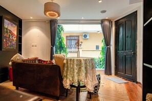 Una sala de estar con un sofá, una mesa con mantel floreado, una lámpara colgante y vistas a un espacio exterior con plantas.