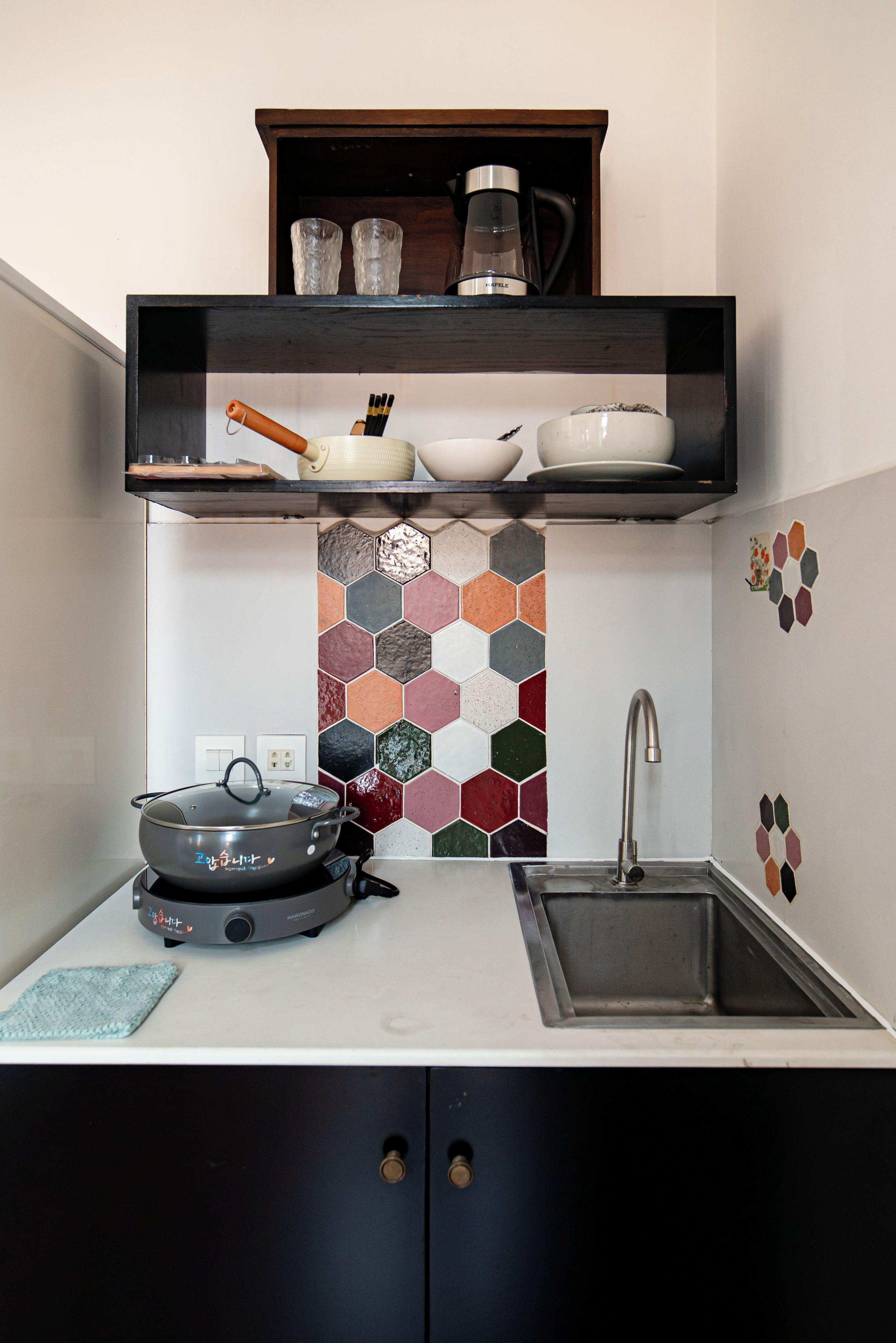 A kitchen with a stove, sink, and a shelf above containing glassware and a kettle.