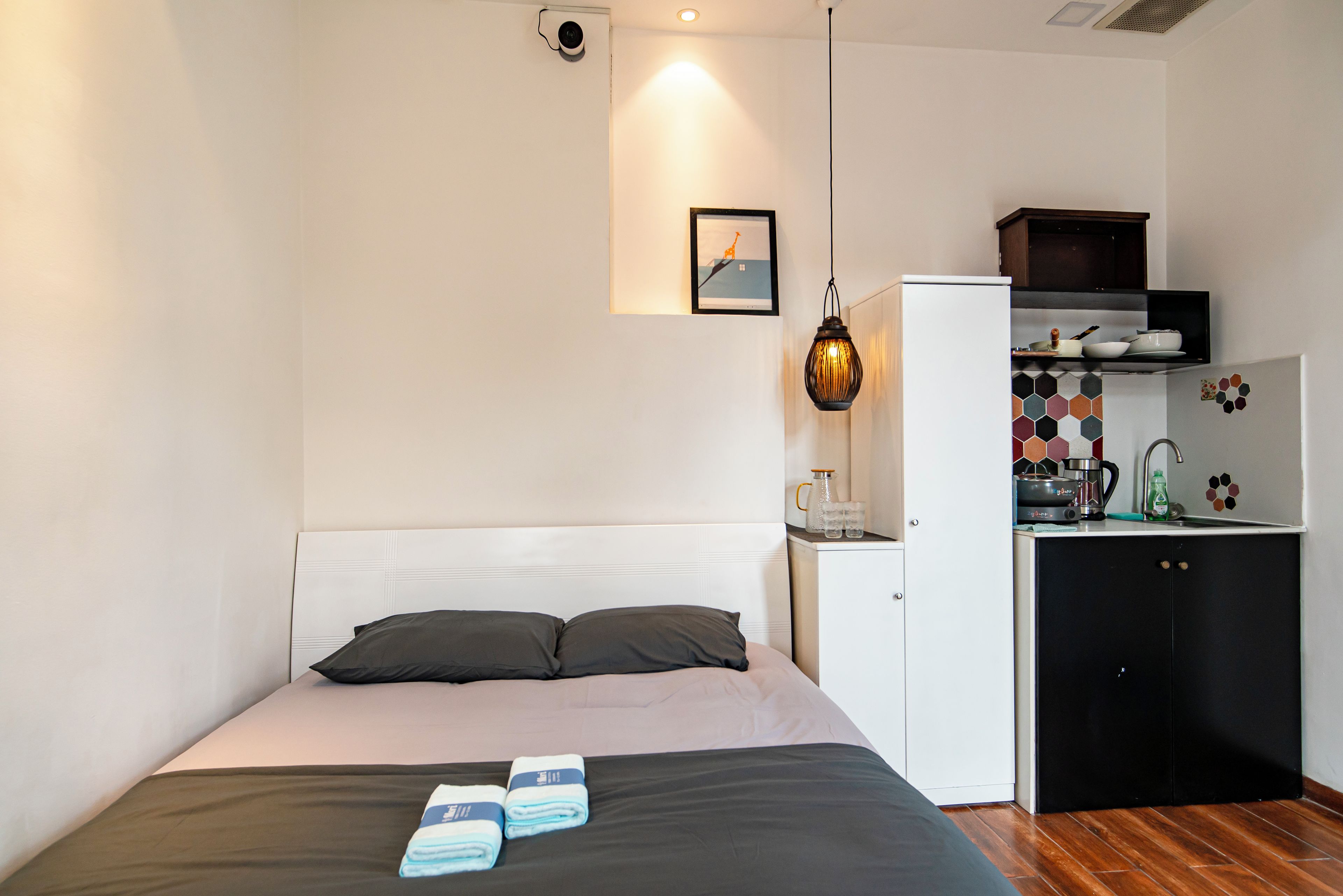 Una camera da letto con un letto, cuscini, un comodino, un angolo cottura e una lampada a sospensione.