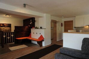 A modern living space with a wooden bench, a grey armchair, and a kitchen area with wooden cabinets. - L-XL up to 12 people (ahrntal)