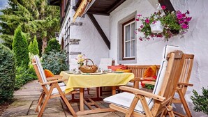 An outdoor dining area with wooden chairs and a table set with a yellow tablecloth, surrounded by greenery and a white building with a window and flower basket. - THE COUNTRY VILLA || Garmisch-Partenkirchen (Garmisch-Partenkirchen)