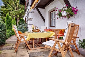 Een eethoek buiten met houten stoelen en een tafel met een gele tafelkleed, omringd door groen en een wit gebouw met een raam en bloemenmand.