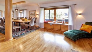 A dining area with a wooden table and chairs, a large window with curtains, and a green sofa with cushions. - THE COUNTRY VILLA || Garmisch-Partenkirchen (Garmisch-Partenkirchen)