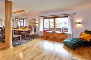 Een eetgedeelte met een houten tafel en stoelen, een groot raam met gordijnen en een groene bank met kussens.