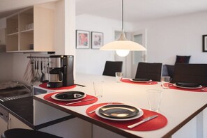 Een eethoek met een witte tafel gedekt voor twee personen, rode placematen en een koffiezetapparaat op het aanrecht.