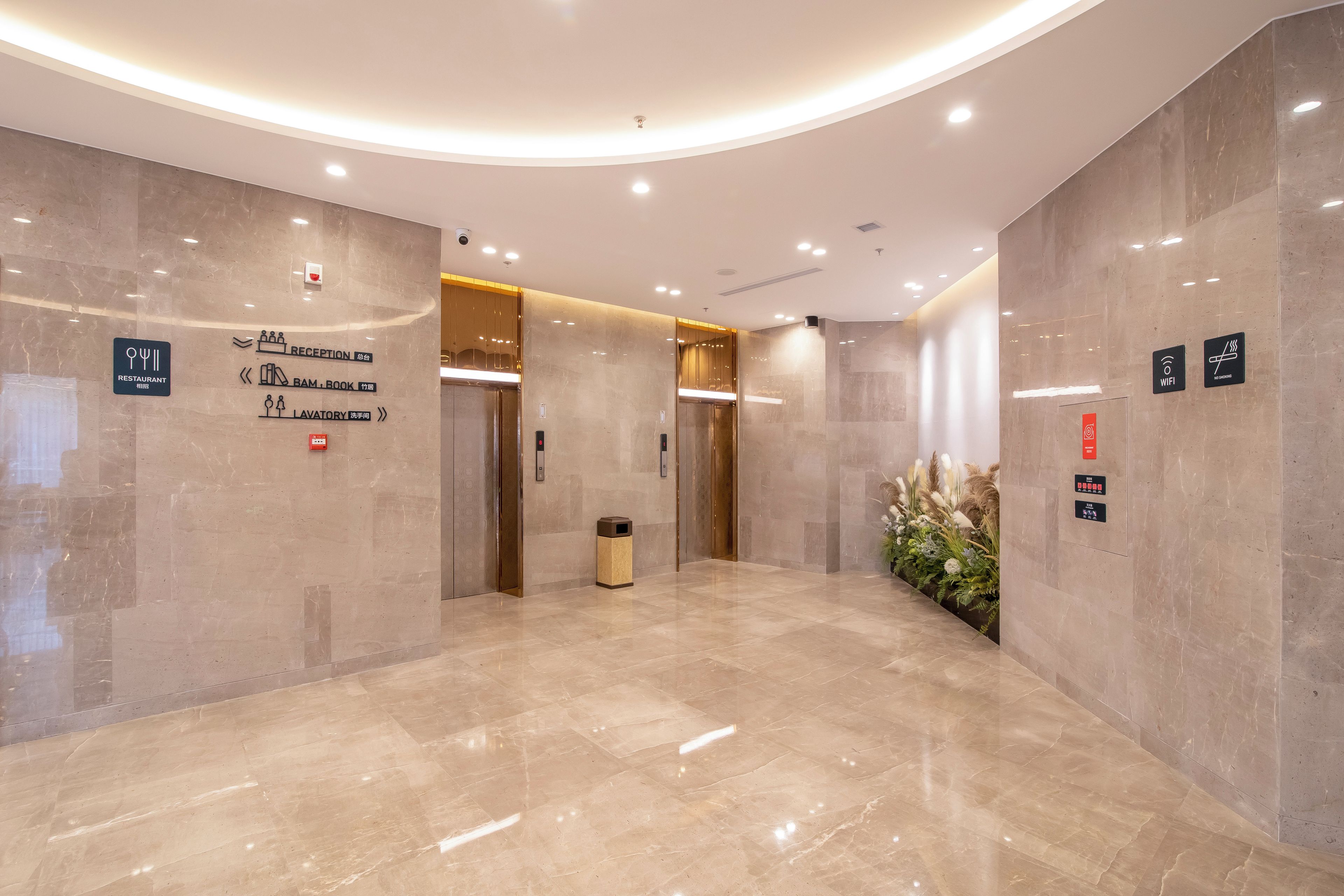Un hall d’entrée moderne avec un sol en marbre, un comptoir de réception et des panneaux indicateurs pour signaler les différents services, tels qu’un restaurant, des toilettes et un ascenseur.