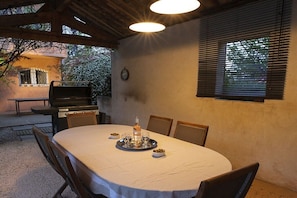 Ein Essbereich im Freien mit einem runden Tisch, Stühlen, einem Grill und einem Fenster mit Rollläden.