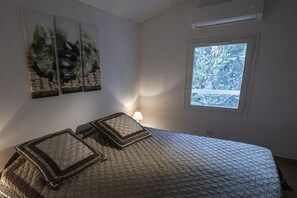Ein Schlafzimmer mit einem Bett, zwei Kissen, einem Nachttisch, einer Lampe, einem Fenster mit Blick auf Bäume und einer Wand-Klimaanlage.