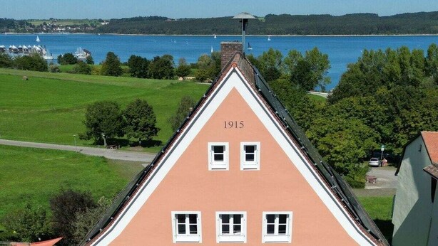 Un bâtiment doté d’un toit triangulaire et portant l’inscription « 1915 », surplombant une étendue d’eau où se trouvent des bateaux.