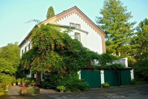 A white building with a green gate, surrounded by lush greenery and flowering plants. - Holiday home in the Palatinate with 1 bedroom (Germersheim)