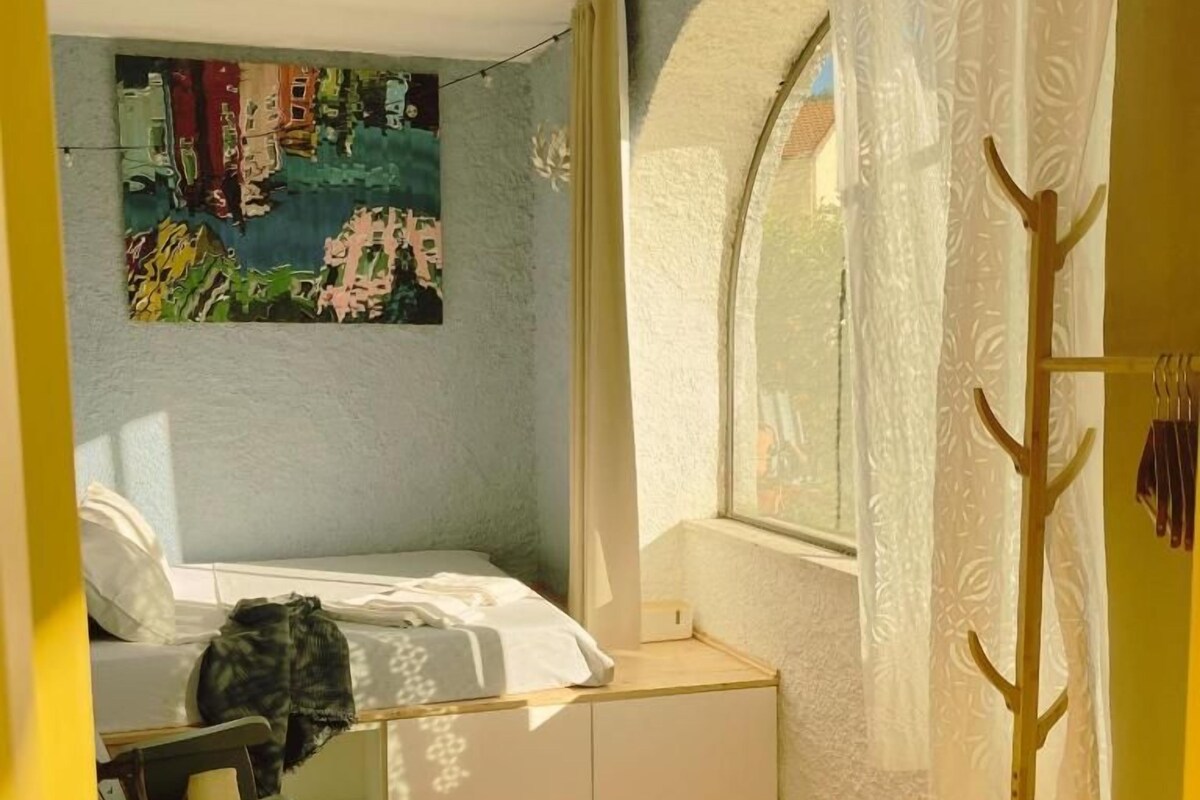 A bedroom with a bed, a window with curtains, a ceiling fan, and a wall-mounted artwork.